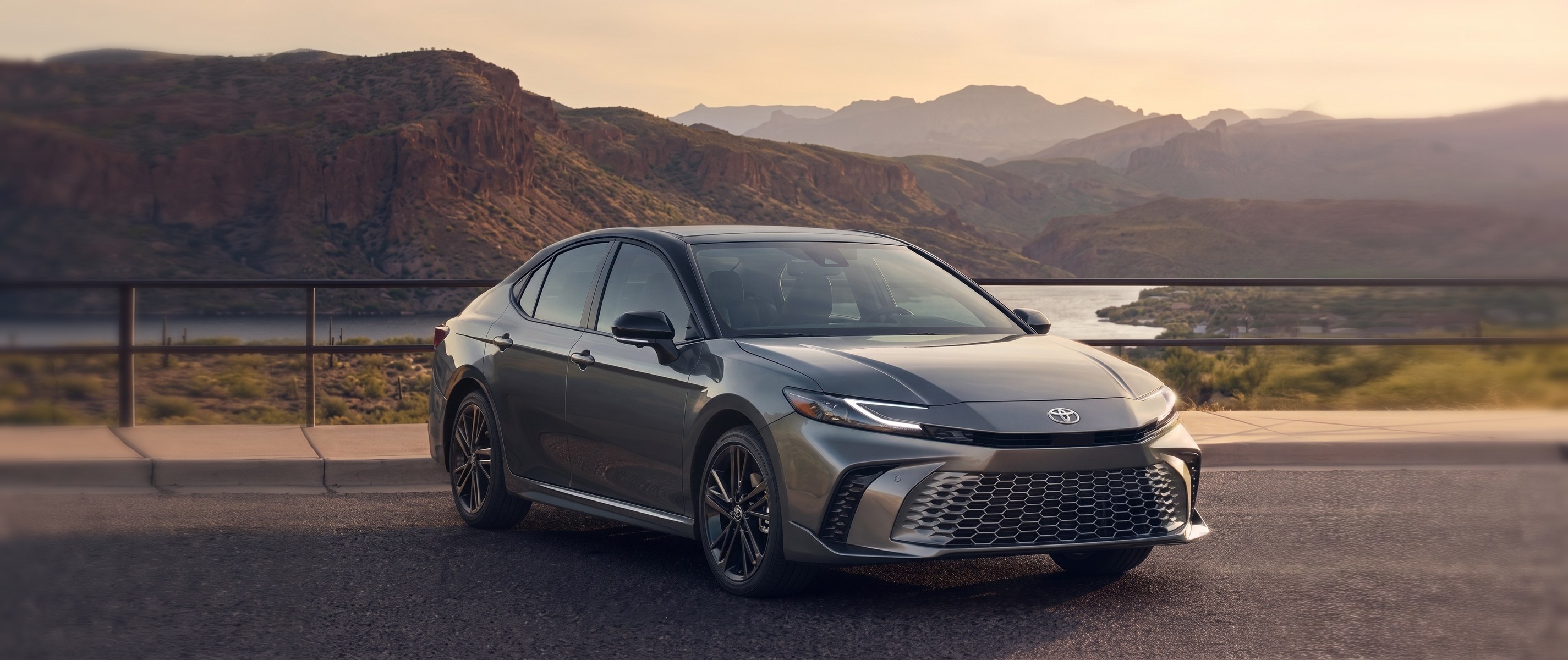 A gray Toyota Camry parked at a scenic overlook at sunset with mountains and a lake behind it